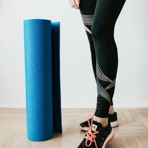 A woman in leggings and sneakers standing with a yoga mat indoors.